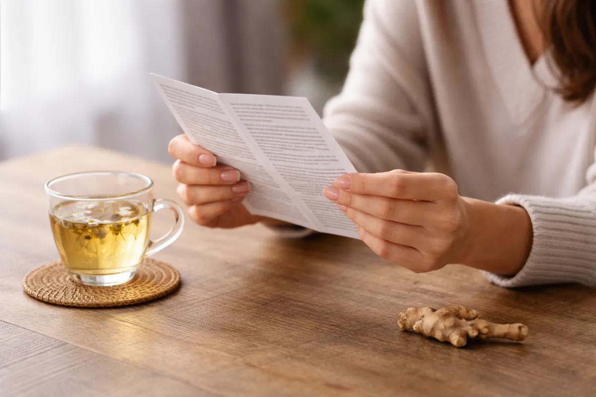 Mains d'une femme tenant une notice dépliée à côté d'une tasse de tisane et d'une racine séchée posée sur une table en bois